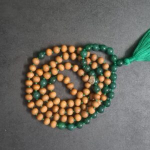 A Green Onyx and Sandalwood Mala Necklace made up of alternating wooden and green beads, with a green tassel at one end, lays coiled on a dark surface.