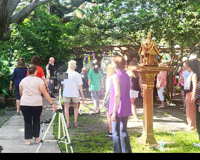 A group of people are gathered outdoors in a garden, participating in an activity. Some are standing while others appear to be listening. There is a decorative structure and colorful flags in the background.