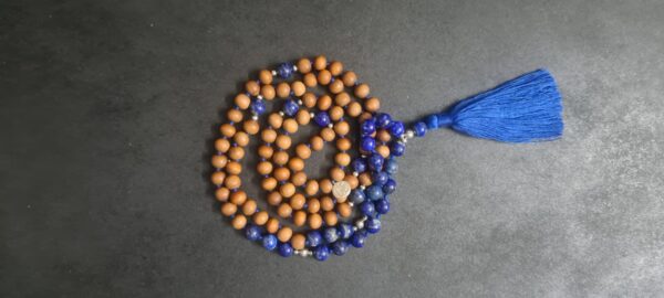 A Blue "Sky" Lapis Lazuli and Sandalwood Mala Necklace with a blue tassel is coiled on a dark surface.