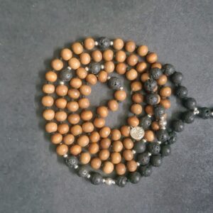 A string of brown and black Grounding Lava Rock and Sandalwood Mala Necklace arranged in a coil on a dark surface with a green tassel at one end.