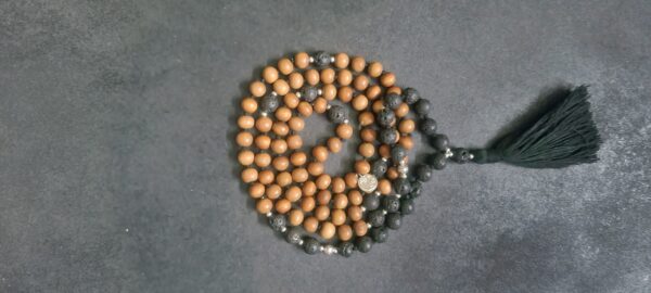 A string of brown and black Grounding Lava Rock and Sandalwood Mala Necklace arranged in a coil on a dark surface with a green tassel at one end.