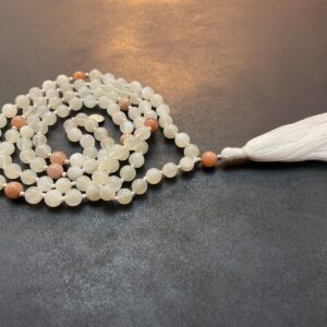 A Moonstone Mala Necklace with a white tassel is laid out on a dark surface.