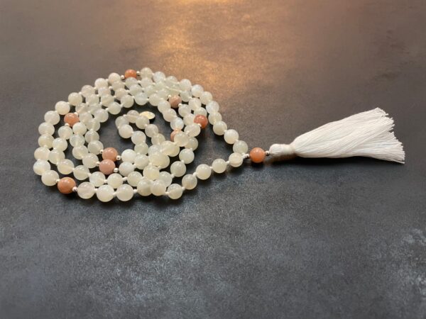 A Moonstone Mala Necklace with a white tassel is laid out on a dark surface.