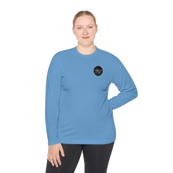 Person wearing a Unisex Lightweight Long Sleeve Tee with a black circle logo on the left chest and black pants, standing with one hand on their hip.