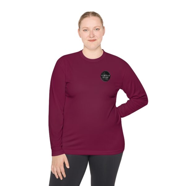 A person wearing a Unisex Lightweight Long Sleeve Tee with a circular logo on the chest and black leggings stands with one hand on their hip against a white background.