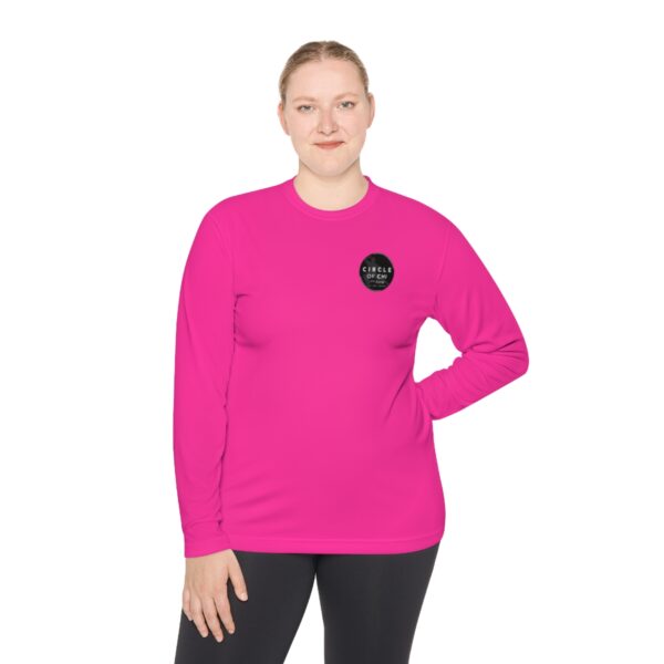 A person wearing a bright pink Unisex Lightweight Long Sleeve Tee with a black circular logo on the left chest and black pants, standing with one hand on their hip.