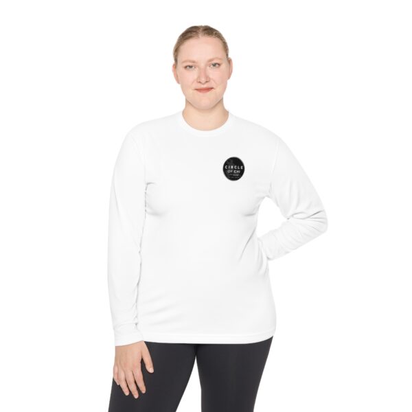 A person wearing a white Unisex Lightweight Long Sleeve Tee with a black circular logo on the left chest area and black pants, posing with one hand on hip.