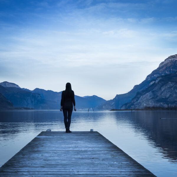 A person stands at the end of a wooden dock, looking out at a calm lake surrounded by mountains under a clear sky, with Music for Spiritual Transformation Vol. 23 No. 2 playing in the background.