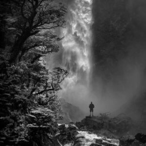 A person stands on rocks near a powerful waterfall, surrounded by lush trees and mist, holding a copy of Music for Spiritual Transformation Vol. 23 No. 3.