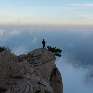 A person stands on the edge of a rocky cliff, overlooking a foggy landscape with the horizon visible in the background, holding a copy of Music For Spiritual Transformation Vol. 23 No. 4.