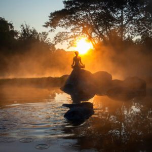 A person meditating on rocks in the middle of a misty pond at sunrise, with trees silhouetted in the background, holding a copy of Music For Spiritual Transformation Vol. 23 No. 5.