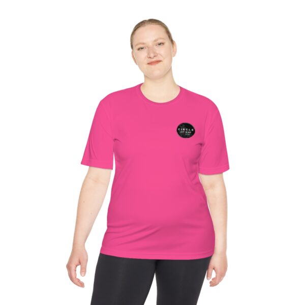 Person standing and smiling, wearing a bright pink Circle of Chi Athletic Tee and black pants. The shirt has a small black circular logo on the left chest area. The background is plain white.