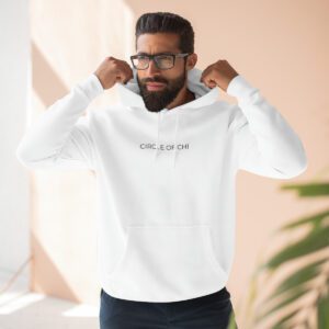 A man with glasses adjusts the hood of his white Three-Panel Fleece Hoodie which has "Circle of Chi" printed on it. He stands indoors, with natural light coming from the left and a plant visible in the foreground.