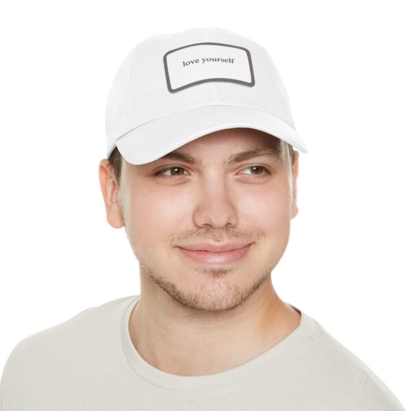 A man wearing a white Love Yourself Dad Hat and a light gray shirt, smiles while looking off to the side.