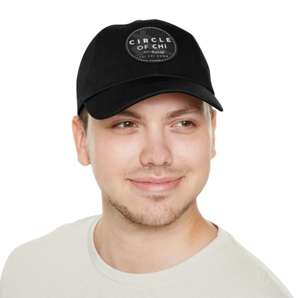 A person wearing a black Circle of Chi Dad Hat with Leather Patch, smiling and looking to the side, dressed in a plain light-colored shirt.