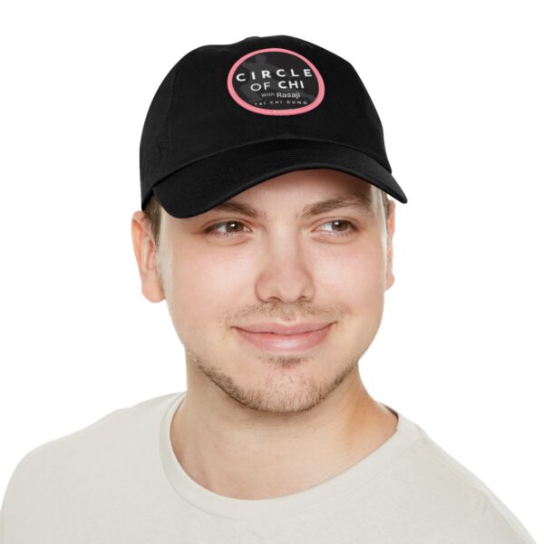 Person wearing a black cap with the text "Circle of Chi Dad Hat with Leather Patch" and a red circular logo, smiling and dressed in a light-colored t-shirt.