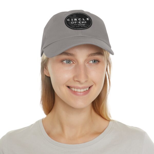 A person with long hair smiles while wearing a Circle of Chi Dad Hat with Leather Patch and a light grey shirt.