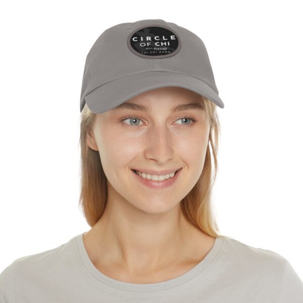 A person with long hair is wearing a gray Circle of Chi Dad Hat with Leather Patch and a gray shirt. They are smiling and looking slightly to the right.