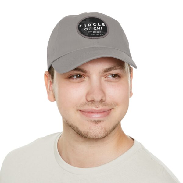 A person wearing a gray cap with the text "Circle of Chi Dad Hat with Leather Patch" on the front, smiling and looking slightly to the side.