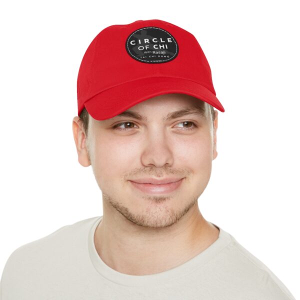 A person wearing a red Circle of Chi Dad Hat with Leather Patch, smiling slightly and looking off to the side. They are wearing a light-colored shirt.