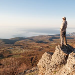 A person stands on a rocky edge, overlooking a vast landscape of rolling hills and distant water under a clear sky, with Music For Spiritual Transformation Vol. 23 No. 6 playing softly in the background.