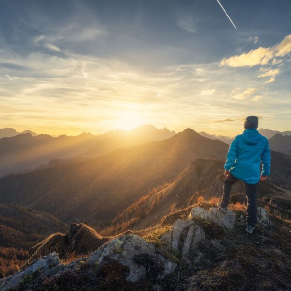 A person in a blue jacket stands on a rocky mountain peak, watching a sunrise over a range of misty mountains with Music For Spiritual Transformation Vol. 24 No. 2 in the background under a clear sky above.