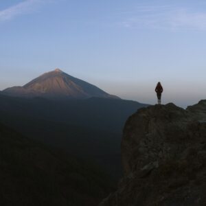 Person standing on a rocky cliff during dawn, overlooking a distant mountain peak under a clear sky, holding Music For Spiritual Transformation Vol. 24 No. 1.