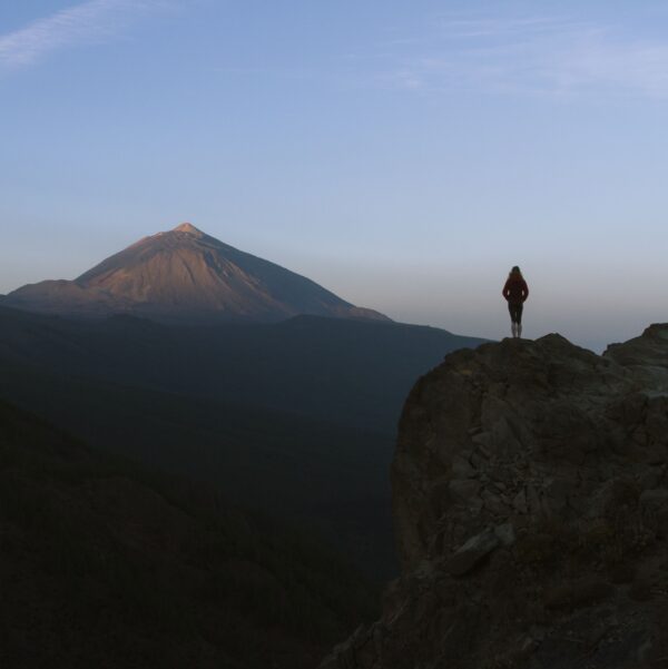 Person standing on a rocky cliff during dawn, overlooking a distant mountain peak under a clear sky, holding Music For Spiritual Transformation Vol. 24 No. 1.